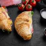 Croissant sandwich with prosciutto, tomatoes, cheese set, with herbs and ingredients, on black stone background